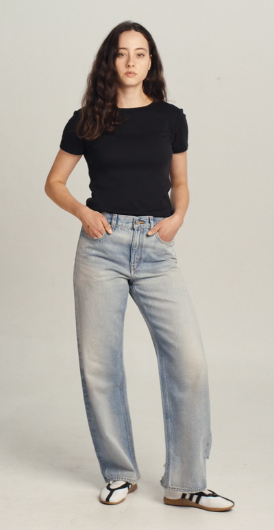 Video of a woman wearing a black wool tee shirt and blue jeans walking into frame, turning to show the tee, then walking out of frame.
