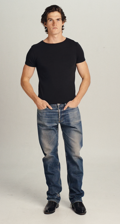 Video of a man wearing a black wool tee shirt and blue jeans walking into frame, turning to show the tee, then walking out of frame.